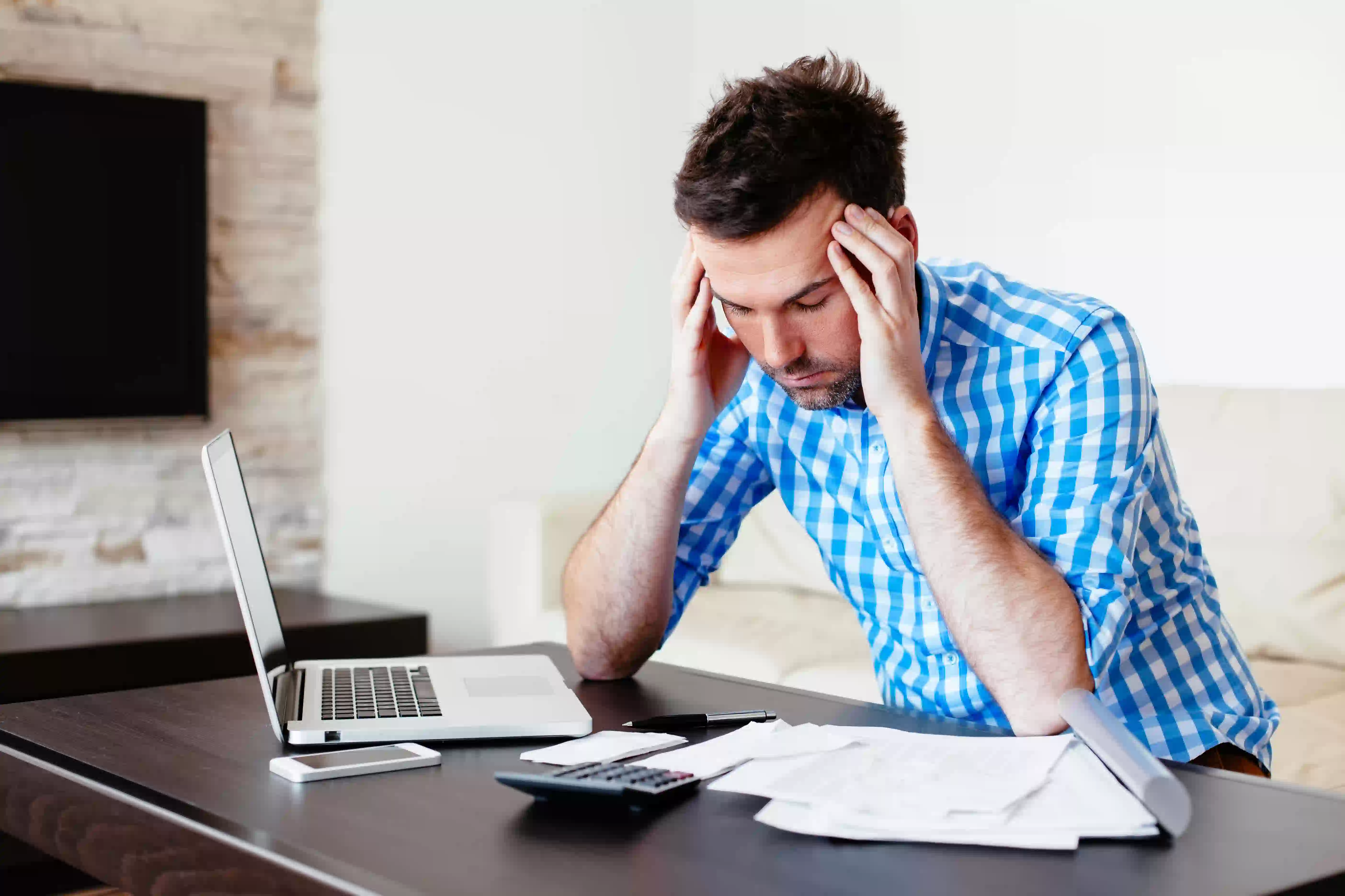 man holding his head in front of laptop, calculator, papers, cellphone